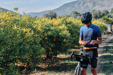 A fit male cyclist standing in an orange garden with trees in the background, cleaning an orange....