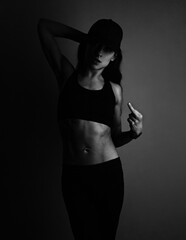Fuck you. Sport sexy muscular woman posing in black sport bra, cap and showing the fuck sign the hand, standing on dark shadow studio background. Front body view. - 737570932
