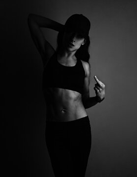 Fuck you. Sport sexy muscular woman posing in black sport bra, cap and showing the fuck sign the hand, standing on dark shadow studio background. Front body view.
