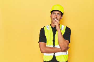 Latino worker in yellow vest and helmet relaxed thinking about something looking at a copy space.