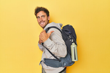 Young Latino with mountain backpack on yellow points with thumb finger away, laughing and carefree.