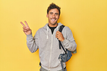 Young Latino with mountain backpack on yellow joyful and carefree showing a peace symbol with...