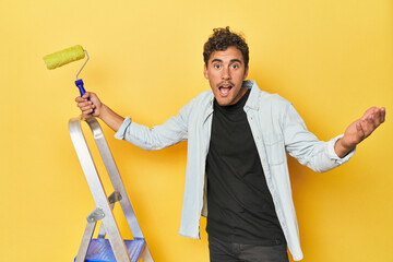 Man with ladder and paint roller on yellow receiving a pleasant surprise, excited and raising hands.