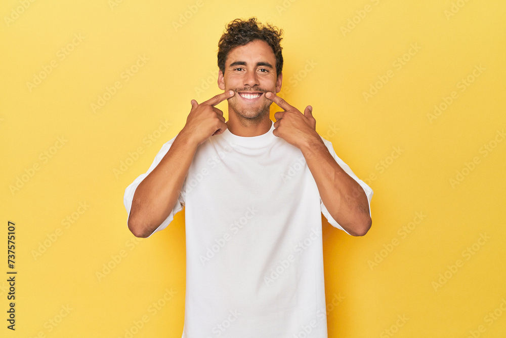 Wall mural Young Latino man posing on yellow background smiles, pointing fingers at mouth. - Wall murals