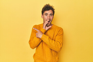 Young Latino man posing on yellow background saying a gossip, pointing to side reporting something.