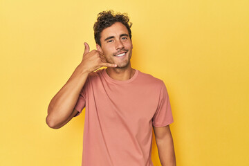 Young Latino man posing on yellow background showing a mobile phone call gesture with fingers.