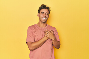 Young Latino man posing on yellow background laughing keeping hands on heart, concept of happiness.