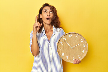 Middle aged woman holding a wall clock on a yellow backdrop pointing upside with opened mouth.