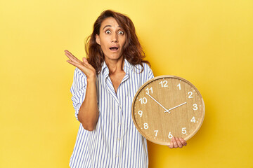 Middle aged woman holding a wall clock on a yellow backdrop surprised and shocked.