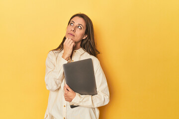 Middle-aged woman with laptop on yellow looking sideways with doubtful and skeptical expression.