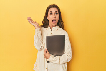 Middle-aged woman with laptop on yellow surprised and shocked.