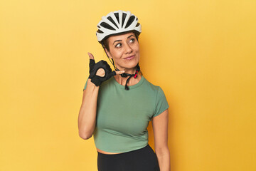 Cyclist middle aged woman with helmet and gloves posed showing a mobile phone call gesture with fingers.