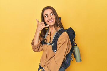 Middle aged woman prepped for hiking, yellow studio shot showing a mobile phone call gesture with fingers.