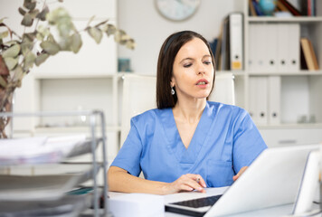 Portrait of female doctor in surgical scrubs sitting at working table in her office and using...