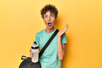 Sportswoman with gym bag & water on yellow studio surprised and shocked.