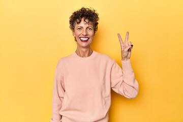 Mid-aged caucasian woman on vibrant yellow joyful and carefree showing a peace symbol with fingers.