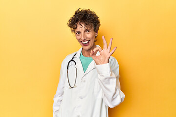 Caucasian mid-age female doctor on yellow studio cheerful and confident showing ok gesture.