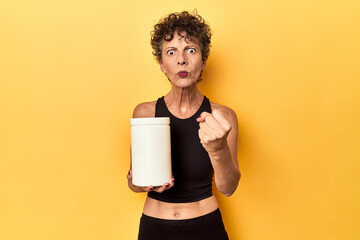 MIddle aged athlete woman holding protein supplement on yellow showing fist to camera, aggressive...