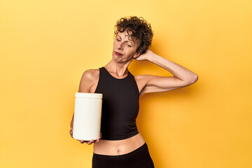 MIddle aged athlete woman holding protein supplement on yellow touching back of head, thinking and...