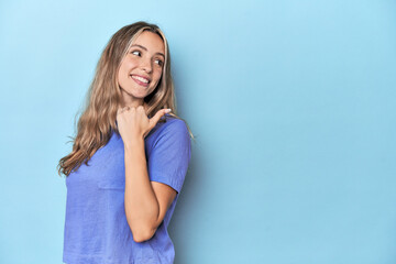 Blonde young caucasian woman in blue studio points with thumb finger away, laughing and carefree.