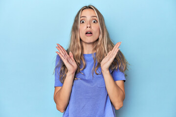 Blonde young caucasian woman in blue studio surprised and shocked.