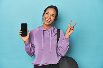 Filipina ready for gym with phone on blue joyful and carefree showing a peace symbol with fingers.