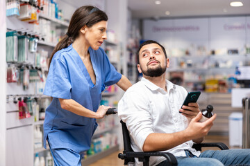 Woman druggist helping man patient on wheelchair to purchase drug in pharmacy. Man using smartphone, browsing information.