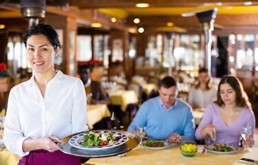 Portrait of positive Asian woman waitress standing in restaurant with dish.