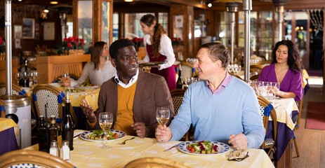 Friendly meeting of two men over dinner with wine in restaurant. Adult European and African...