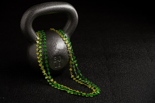 St. Patrick’s Day fitness, silver powder coated iron kettlebell on a gym floor with three strands of green and gold metallic shamrock fun necklaces
