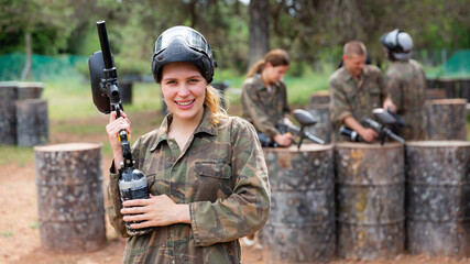 Fototapeta premium Young smiling woman paintball player in camouflage posing with gun outdoors