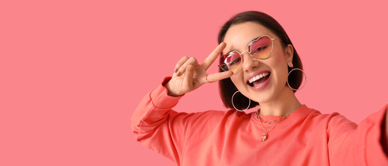 Beautiful young woman in stylish sunglasses showing victory gesture on pink background with space for text