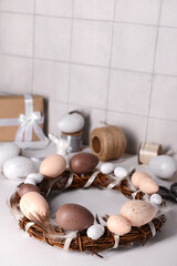 Easter wreath made of eggs and feathers with supplies on table near white wall