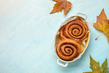 Baking dish of tasty cinnamon rolls and autumn leaves on blue background