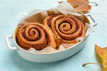 Baking dish of tasty cinnamon rolls and autumn leaves on blue background