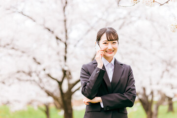 満開の桜の下でスマホの画面を指差すスーツ姿の女性（求人・転職・就活・進学）
