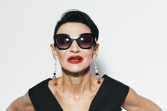 Fashionable woman wearing sunglasses and black dress posing in front of white wall for camera shot