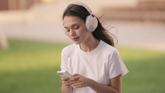 A young woman engages with her smartphone and listens to music using noise-cancelling headphones, exuding a sense of calm and relaxation outside.