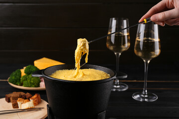 Woman dipping piece of ham into fondue pot with melted cheese at table, closeup