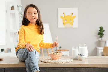 Cute little Asian girl with bowl of cereal rings sitting on counter in kitchen