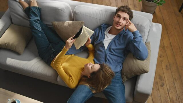 Young spouses reading book comfortable couch top view. Couple talking together
