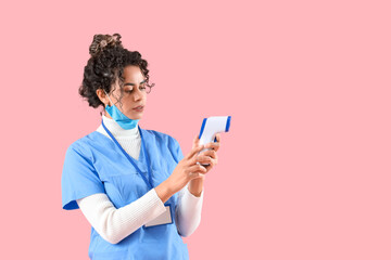 Female African-American medical intern with infrared thermometer on pink background