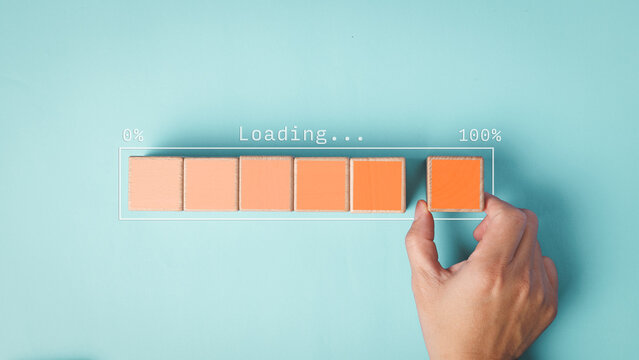 A hand completes a wooden block loading bar to 100 percent on a blue background, visually representing concepts of progress, completion, and achievement.