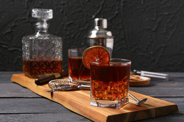 Board with glasses of Old Fashioned Cocktail, dried orange slice and bar tools on wooden table near black wall
