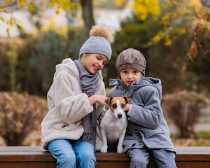 Fototapeta premium Brother and sister sit in an embrace with a dog on a bench for a walk in the autumn park. Boy, girl and jack russell terrier. 