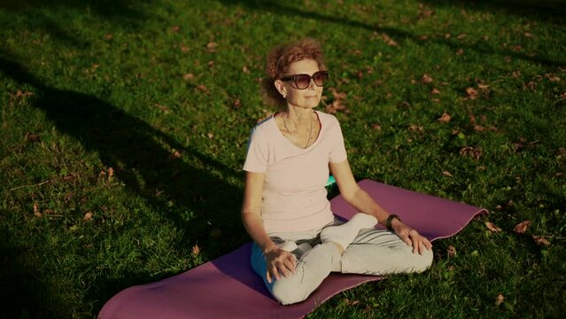 Senior woman meditating and exercising yoga lotus position outdoors. Ederly female doing stretching exercises on yoga mat in park on green lawn at sunset. Feeling energy. Concept calm and meditation.