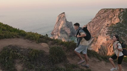 Active young man and woman with backpacks hiking on a coastal trail overlooking the ocean during a beautiful sunset, embodying adventure, travel, and fitness. - Powered by Adobe