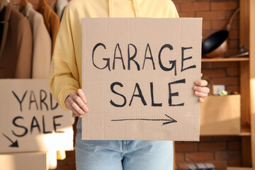 Woman holding cardboard with text GARAGE SALE in room of unwanted stuff