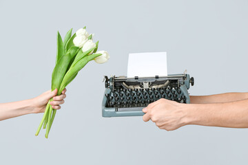 Hands holding vintage typewriter with blank paper and tulips on grey background. Women's Day...