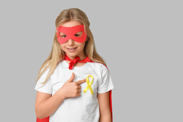 Cute teenage girl in superhero costume pointing at yellow ribbon on grey background. International Childhood Cancer Day